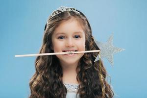 Horizontal shot of attractive smiling small kid holds magic wand in mouth, has brown curly hair, poses over blue background, looks directly at camera, wears crown. Children and magic concept photo
