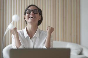 Excited italian lady clenching fists with joy while sitting in front of laptop at workplace photo
