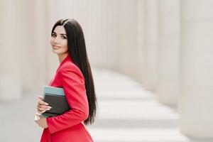 Sideways shot of elegant brunette lady with dark long straight hair, dressed in fashionable red suit, has manicure, carries diary, stands outdoor over white building. Female employee in formal apparel photo