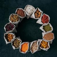 Assorted legumes and beans in sacks standing in circle, isolated over dark background with copy space in middle. Healthy eating concept