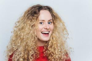 Una foto de una hermosa mujer satisfecha con una expresión agradable y alegre, con el pelo rizado y tupido, vestida con un suéter rojo, no espera escuchar algo emocionado, aislada sobre fondo blanco.