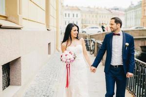 Attractive female with dark hair, wears white dress, holds bouquet, keeps bridegrooms hand, look at each other with great love, have walk across ancient city on bridge. Positive emotions concept photo