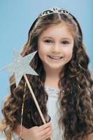 Cute little girl with positive expression, holds wand, has curly dark hair, carries wand, believes in fairy tale, isolated over blue background, wishes dreams come true. Children and magic concept photo