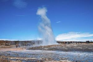 Tourists enjoying view of Strokkur geyser eruption in valley against blue sky photo