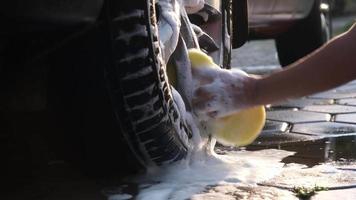 Nahaufnahme der Hand einer Frau, die die Leichtmetallräder eines Autos mit Schwamm und Seife auf der Autowaschanlage wäscht. manuelle autowäsche mit weißer seife. autowaschservice-konzept. video