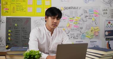 retrato de un feliz hombre de negocios asiático escribiendo una laptop y mirando la cámara mientras se sienta en el escritorio del lugar de trabajo en la oficina del hogar. trabajador de sexo masculino sonriente que trabaja en el ordenador portátil. video