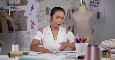 Asian woman fashion designer looking through color swatches or checking data in paper. Focused female clothing designer working while sitting at desk in her office.