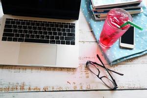 portátil con gafas, teléfono inteligente, un vaso de bebida fría y una revista sobre una mesa de madera, trabajando de forma remota. foto