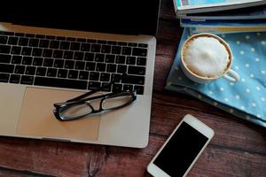 portátil con gafas, teléfono inteligente, una taza de café caliente y una revista sobre una mesa de madera, trabajando de forma remota. foto