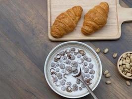 Top view of breakfast cereal with milk, croissants and pistachios on wooden background with copy space. photo