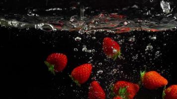 Slow motion strawberries dipped falling in water, bubbles and drops on black background close up video