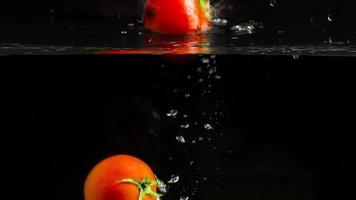 Slow motion many tomatoes dipped falling in water, bubbles and drops on black background close up