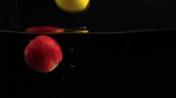 Slow motion many fruits dipped falling in water, bubbles and drops on black background close up video