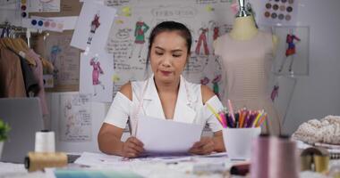 Asian woman fashion designer looking through color swatches or checking data in paper. Focused female clothing designer working while sitting at desk in her office. video