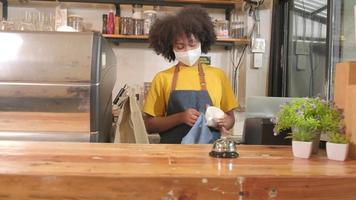 la barista afroamericana trabaja limpiando la taza de café, mirando a través de la ventana del café, esperando a los clientes en un nuevo servicio de estilo de vida normal, el impacto comercial de la cuarentena pandémica covid-19.