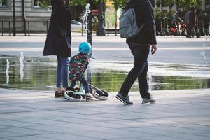 Little kid with small bicycle with two parents walking in early summer on paved city square with wet ground near fountains photo