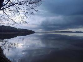 Heavy rain cloud moving over the lake with clean clear water and visible plank lying in it and leafless tree branches in spring photo