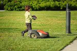 Vilnius, Lithuania. June 06 2022 - Little boy having fun running with automated lawnmower cutting grass in a park photo