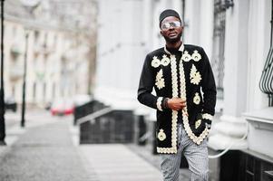 hombre africano mega elegante en pose de chaqueta tradicional. chico negro de moda con sombrero y gafas de sol con cigarro en la mano. foto