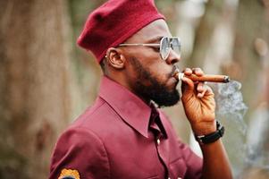 Portrait of African American military man in red uniform, sungalasses and beret. Captain smoke cigar. photo