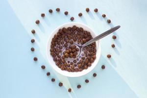 Chocolate cereal balls in white bowl on blue background. Heart shape from flakes. Dry breakfast cereal top view. Concept for a healthy diet photo