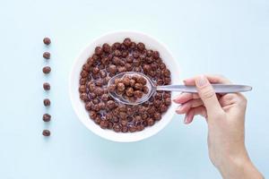 Hand with a spoon over a bowl with chocolate cereal balls on blue background.Dry breakfast cereal top view. Concept for a healthy diet photo