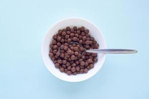 bolas de cereal de chocolate en un tazón blanco sobre fondo azul. vista superior de cereal de desayuno seco. concepto para una dieta saludable foto