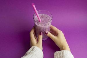 Female hands holds glass of berry smoothie with straw on purple background. Pink milkshake. Healthy drink. Blueberry cocktail photo