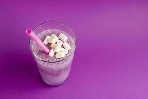 Glass of berry smoothie and marshmallow with straw on purple background. Pink milkshake with copy space. Healthy drink. Blueberry cocktail photo