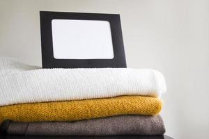 Close-up blank frame and stack of folded clothes on white background wall. Multicolored knitted textiles. Concept with copy space for text photo