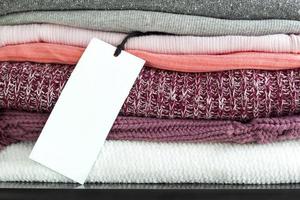 Label white price tag on stack of folded colorful clothes. Mockup label on background of knitted sweaters for design and branding. Copy space for text photo