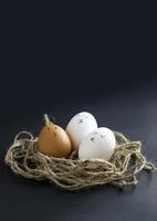 Nest with fresh chicken egg with drawn faces on black paper background. Concept for Easter with copy space. Creative layout made of white and brown eggs photo