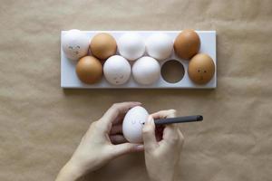 Hands paint cute face on chicken eggs on a brown paper background. Concept for Easter with copy space. Creative layout made of white and brown eggs. Top view photo