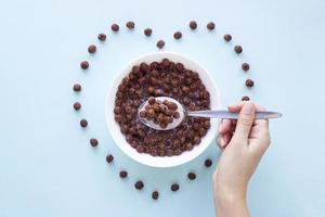 Hand with a spoon over a bowl with chocolate cereal balls on blue background.Dry breakfast cereal top view. Heart shape from flakes. Concept for a healthy diet photo