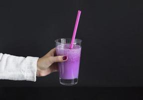 Female hand holds glass of berry smoothie with straw on black background. Pink milkshake with copy space. Healthy drink. Blueberry cocktail photo