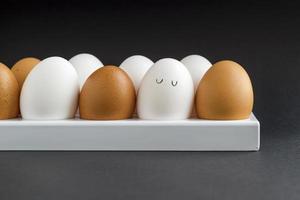 Fresh chicken egg in container on black paper background. Concept for Easter with copy space. White and brown eggs. Painted cute face on a white egg photo