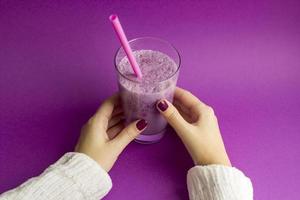 Female hands holds glass of berry smoothie with straw on purple background. Pink milkshake. Healthy drink. Blueberry cocktail photo