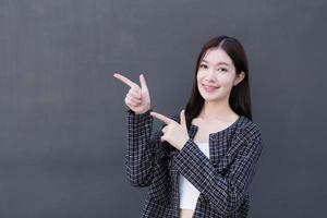 Asian working woman who wears black suit with braces on teeth is pointing hand to present something on the dark gray wall. photo