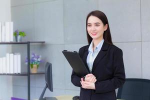 Professional Asian business young woman in black suit smiles happily while she works and holds clipboard in office. photo