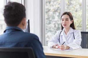 Asian professional  woman doctor who wears medical coat talks with a man patient to consult and suggest healthcare information to him at the examination room in the hospital. photo