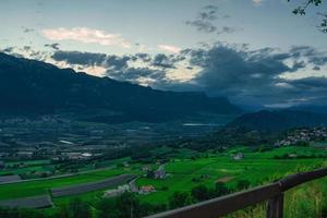 Mountain landscape imaged during sunset under a dramatic sky, dark clouds over mountain with a lake and a rural village in background photo