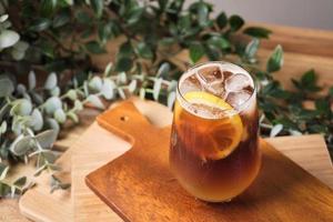 café helado negro mezclado con jugo de limón y rodajas de limón en un vaso con trozos de hielo sobre una mesa de madera, luz natural, bebida fresca de verano por la mañana foto