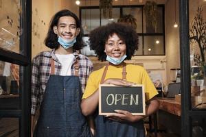 Two young startup barista partners stand at casual cafe door, write and show open sign before taking off face masks, happy and cheerful smiles with coffee shop service jobs after COVID-19 lifestyle. photo