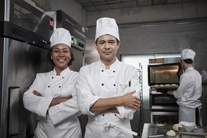 Pastry foods professional partners, two happy chefs team staff in white cooking uniforms stand, arms crossed with confidence, cheerful smiles with commercial culinary jobs in restaurant kitchen. photo