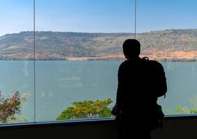 silueta trasera de un turista de mochila parado y mirando malditamente y vistas a la montaña en una ventana transparente desde la habitación interior foto