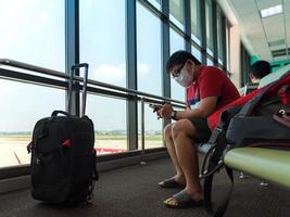 hombre con ropa informal sentado cerca del equipaje usa una máscara protectora mirando seriamente el teléfono inteligente revisando noticias o trabajando en línea durante la espera del vuelo foto