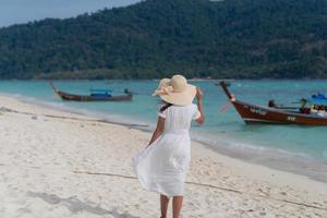 rear view of woman in white dress holding wide brim hat walking on windy blue sea beat with wooden boats photo