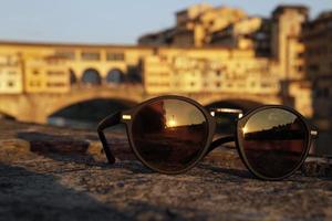 A pair of sunglasses reflects the setting sun with the famous Ponte Vecchio in Florence, Italy, in the background photo