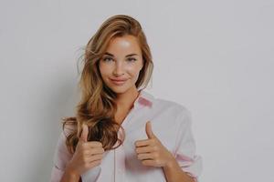 Pretty European woman with satisfied expression recommends product or best service keeps thumbs up photo