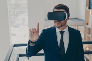 Smiling successful businessman in suit standing in office interior and using VR glasses for work photo